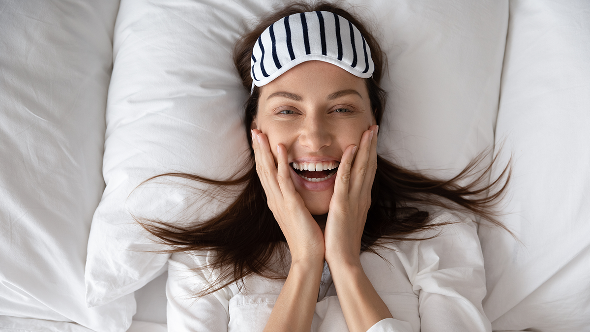 Woman laughing in bed, sleep mask pushed up to her forehead.